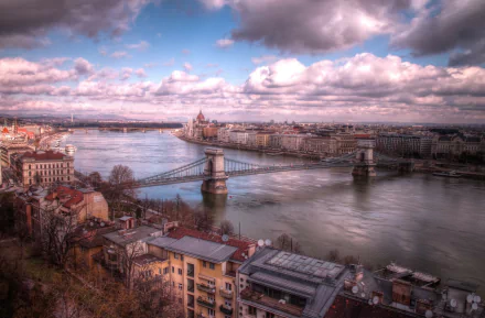 A stunning HD desktop wallpaper of Budapest featuring the iconic man-made Chain Bridge over the Danube River under a dramatic cloudy sky.