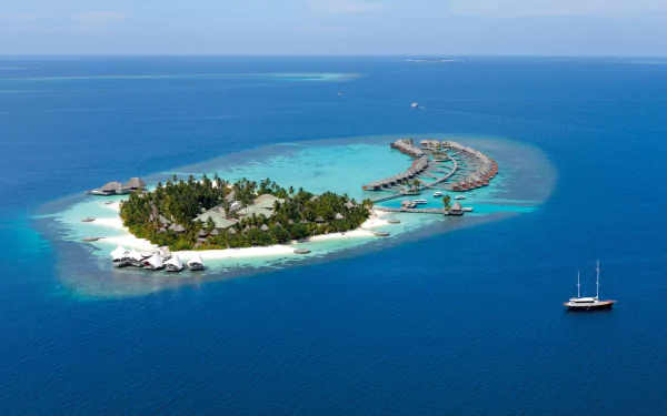 Aerial view of a tropical Maldives atoll with lush village island, overwater hotel bungalows, and a ship sailing in the deep blue sea under clear skies.