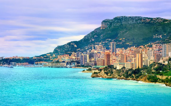 Monaco coastline with turquoise sea, densely packed high-rise buildings at the foot of green hills; man-made skyline against a blue sky — 2K Quad HD PC desktop wallpaper/background.