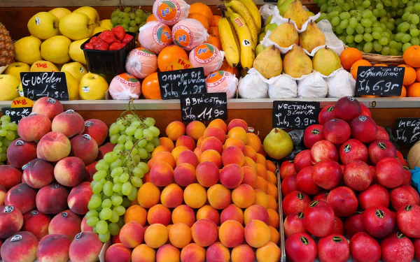 A vibrant display of fresh fruits, including oranges, peaches, bananas, pears, grapes, and apples, showcased at a market stall, with colorful labels and pricing.