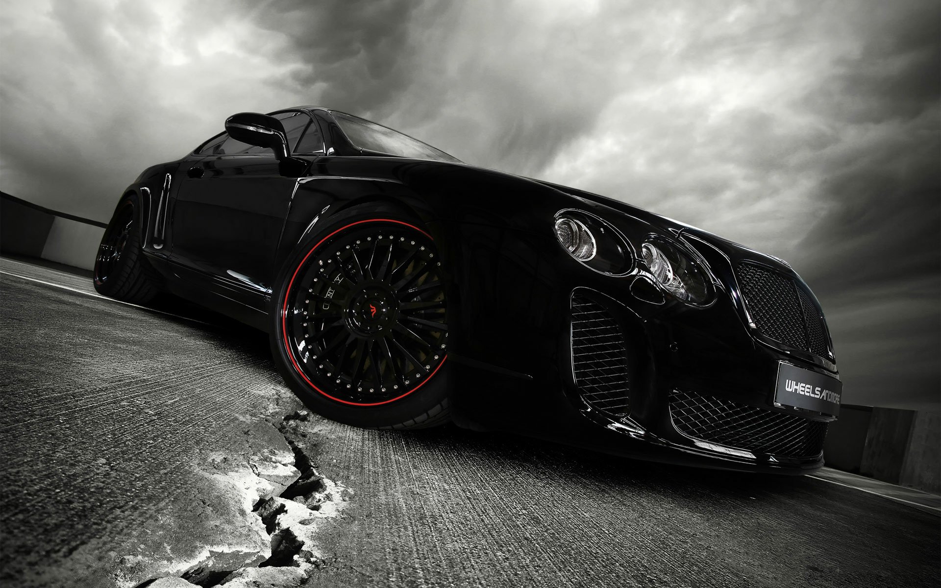 HD desktop wallpaper of a black Bentley Continental coupe with red-accented rims, parked on cracked concrete beneath dramatic stormy clouds.