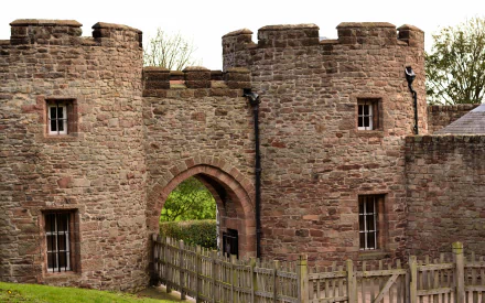 Man-made Beeston Castle stone gatehouse with arched entrance and turrets, framed by a wooden fence and trees — HD PC desktop wallpaper background.