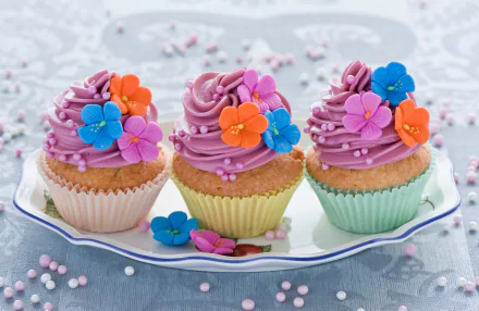 A vibrant HD desktop wallpaper featuring three colorful cupcakes adorned with purple frosting and floral decorations on a decorative plate, surrounded by pastel sprinkles.