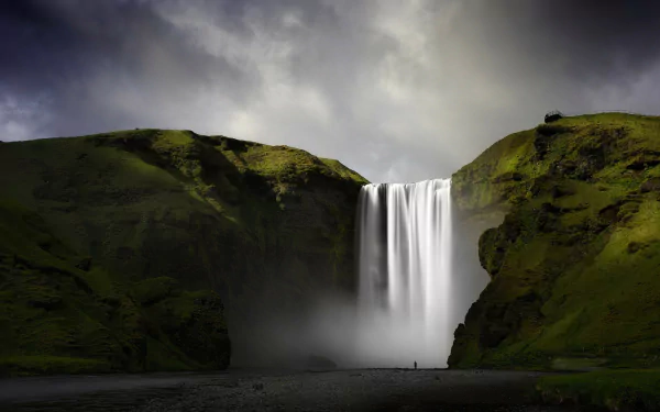 HD desktop wallpaper of Skógafoss waterfall in Iceland, showcasing cascading water surrounded by lush green cliffs under a moody, cloudy sky.