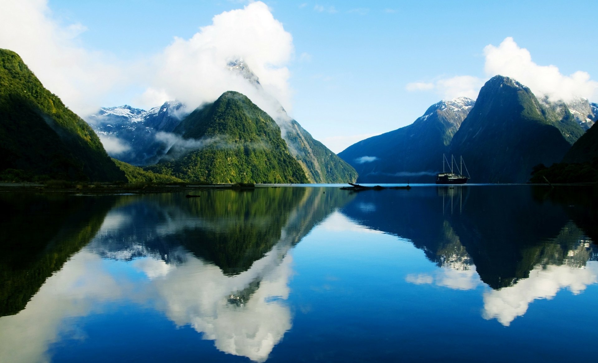 Mitre Peak and surrounding mountains reflected in the calm fjord waters of Milford Sound, New Zealand, under a bright sky with scattered clouds.