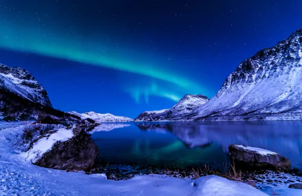 HD wallpaper of a stunning aurora borealis over a snow-covered mountain landscape with a clear starry sky and calm lake reflection.