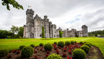 HD desktop wallpaper of Ashford Castle, a majestic man-made stone fortress surrounded by manicured gardens and lush green lawns under a cloudy sky.