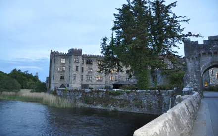 HD PC desktop wallpaper of Ashford Castle, a man-made stone fortress by a lakeside with an arched bridge, turrets and tall trees under a dusky sky.