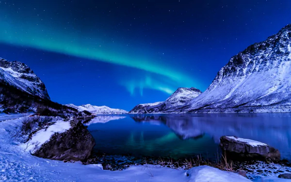 HD wallpaper of a stunning aurora borealis over a snow-covered mountain landscape with a clear starry sky and calm lake reflection.