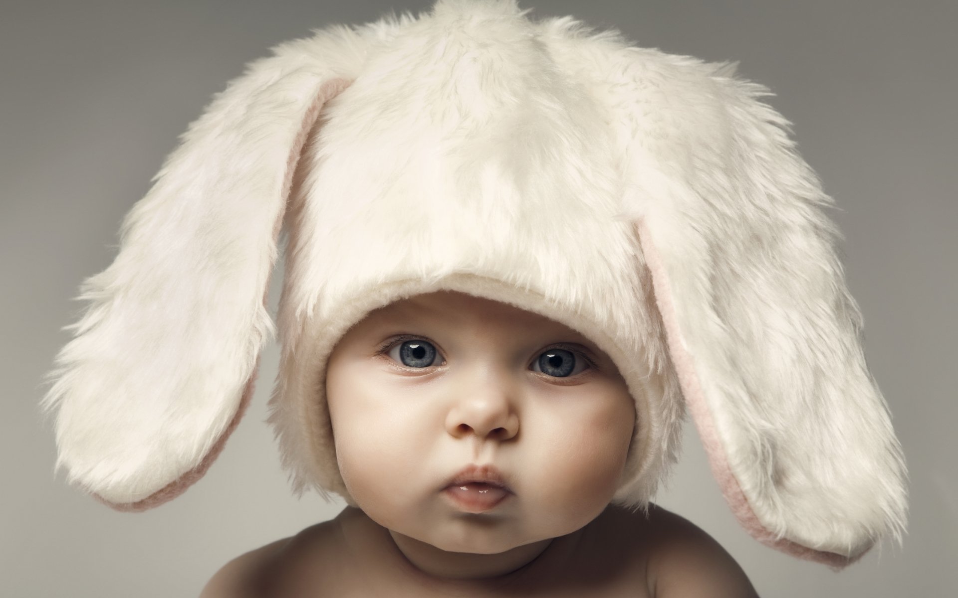 A cute baby with blue eyes wears a fluffy bunny hat, set against a neutral background. This HD image serves as a charming desktop wallpaper.