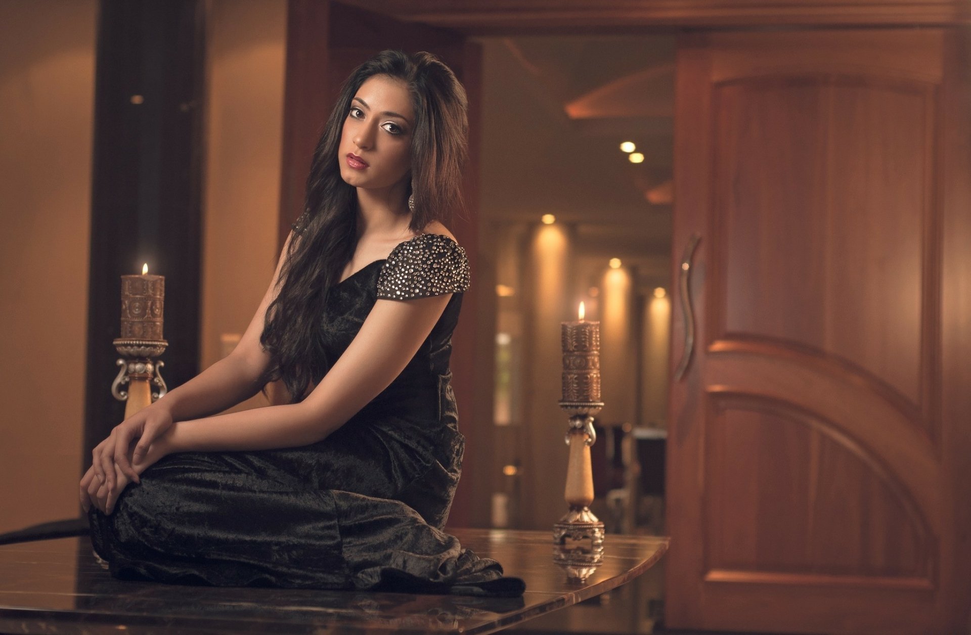 A woman in a black dress sits gracefully on a table in an elegantly lit room, creating a stylish HD desktop wallpaper and background.