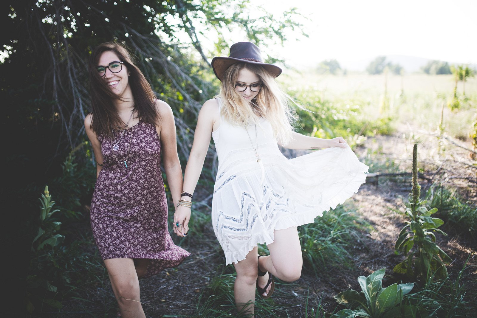 Two women in love holding hands, laughing as they walk through a sunlit meadow; one in a white dress and hat, the other in a patterned dress. HD PC desktop wallpaper/background.