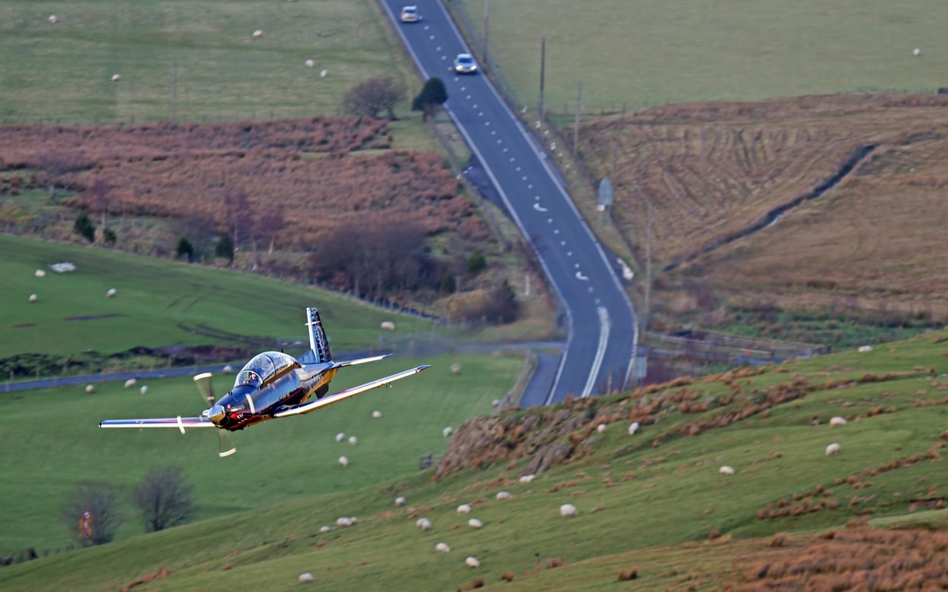 Military Beechcraft T-6 Texan II flying low over green rolling hills beside a winding road — HD PC desktop wallpaper/background.