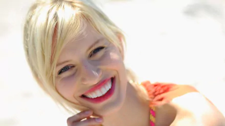 HD desktop wallpaper: close-up of a smiling blonde woman with red lipstick in bright sunlight, beach-style portrait conveying a relaxed, cheerful mood.