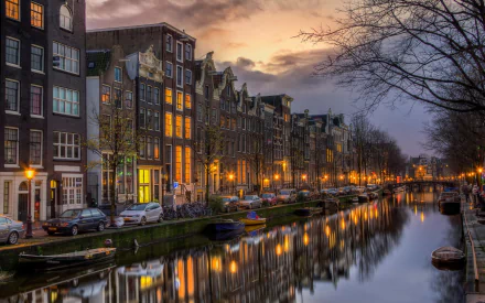 A serene Amsterdam canal at dusk, showcasing illuminated buildings along the water, with trees and parked boats, creating a picturesque night scene. An enchanting HD wallpaper.