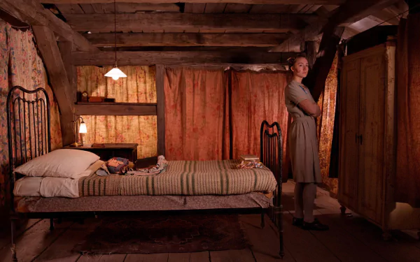 HD PC desktop wallpaper: moody attic bedroom set from the film The Grand Budapest Hotel — iron-framed bed with striped coverlet and a young woman standing by a wooden wardrobe.