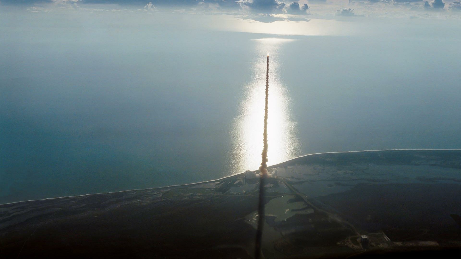 HD PC desktop wallpaper background: space shuttle vehicle ascending over a coastline, its rocket plume forming a straight trail toward a bright sun reflection on the calm ocean.