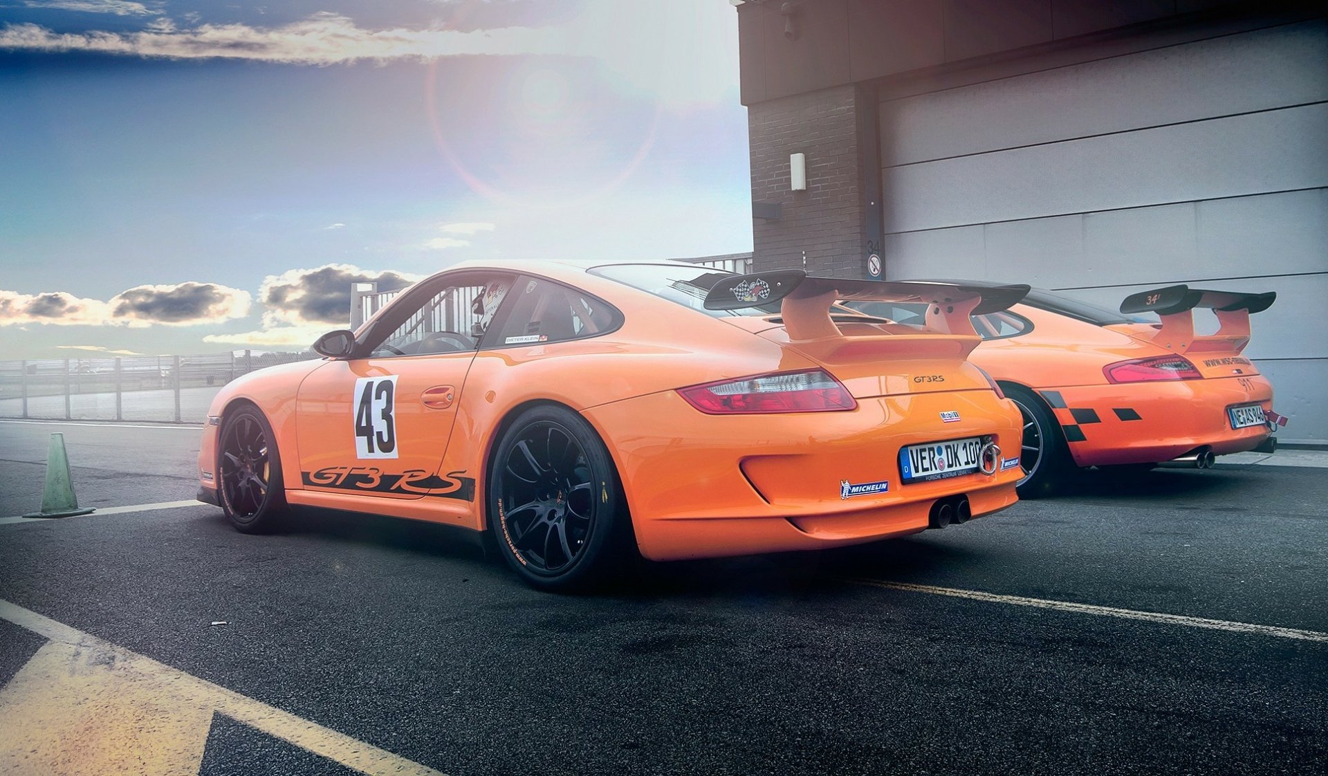 HD PC desktop wallpaper of two orange Porsche 911 GT3 vehicles (car #43) parked in a pit lane with large rear wings under a dramatic sky