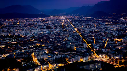 A 4K Ultra HD nighttime aerial view of Grenoble, showcasing glowing city lights and man-made structures against a backdrop of mountains.