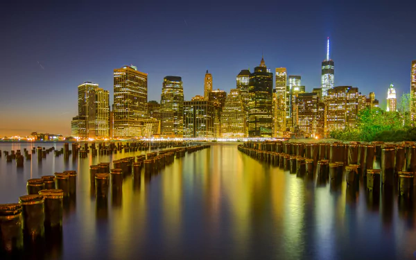 4K Ultra HD nighttime view of Manhattan skyline with illuminated skyscrapers reflecting on water, framed by wooden pilings in the foreground.