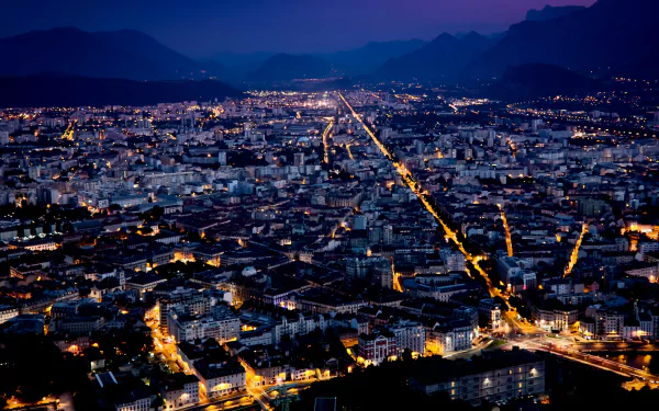 A 4K Ultra HD nighttime aerial view of Grenoble, showcasing glowing city lights and man-made structures against a backdrop of mountains.