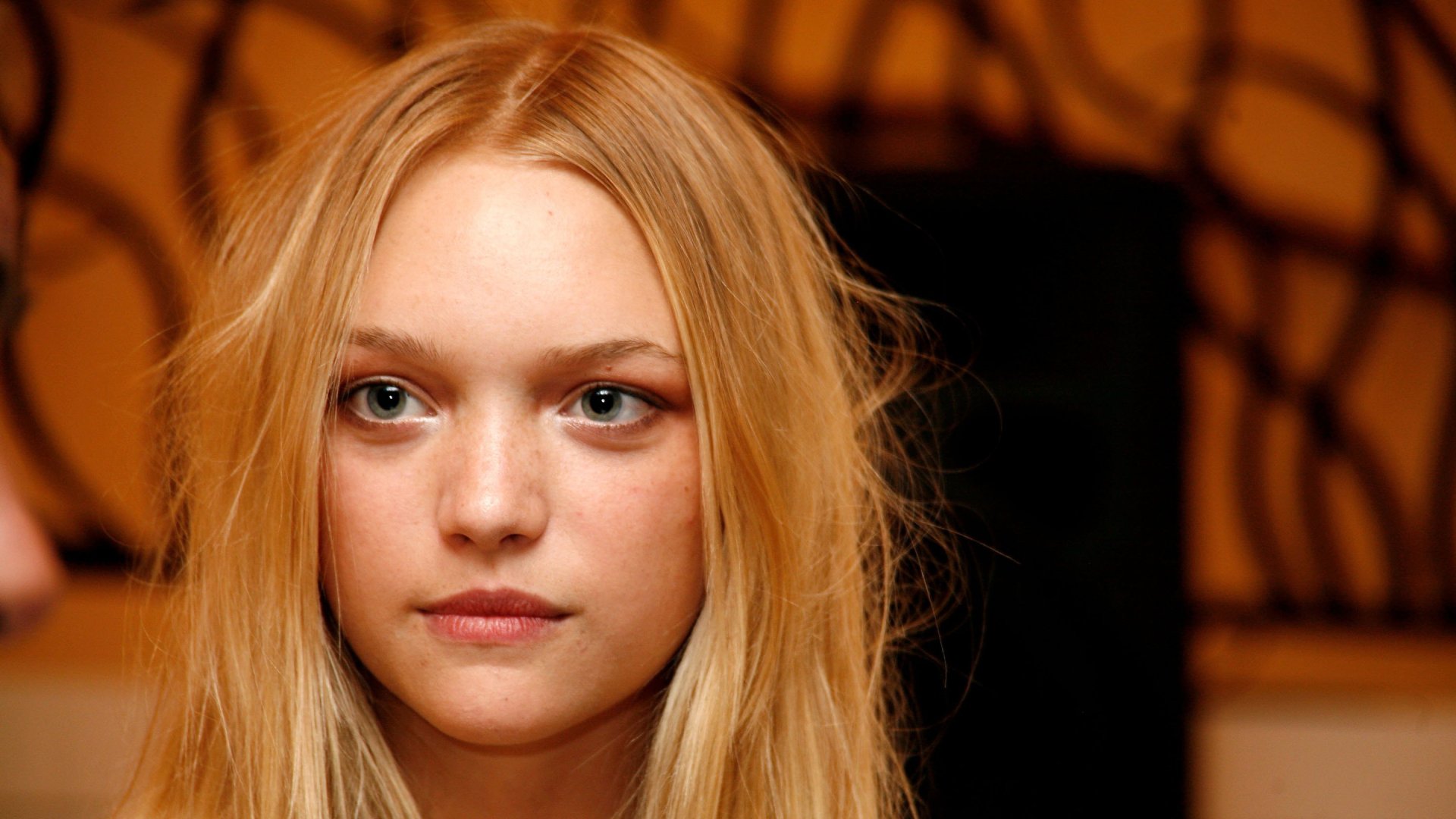 Close-up portrait of a young blonde model with loose hair against a warm patterned backdrop — 2K Quad HD PC desktop wallpaper.