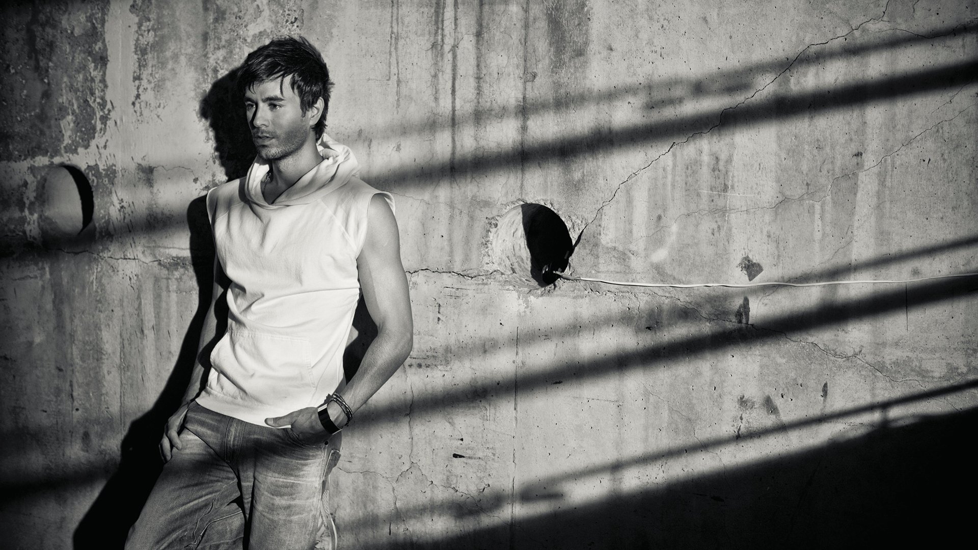 A striking black and white HD wallpaper featuring Enrique Iglesias, standing against a textured concrete wall with dramatic shadows, capturing his confident, artistic persona.