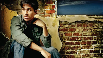 High-definition wallpaper featuring Enrique Iglesias, sitting against a rustic brick wall, exuding a thoughtful expression in a stylish, casual outfit.