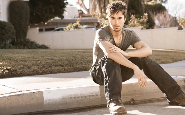 HD desktop wallpaper of Enrique Iglesias sitting on a curb, exuding a relaxed vibe, against a suburban backdrop. The image seamlessly blends elements of music and a serene outdoor setting.