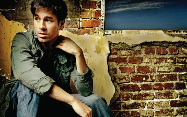 High-definition wallpaper featuring Enrique Iglesias, sitting against a rustic brick wall, exuding a thoughtful expression in a stylish, casual outfit.
