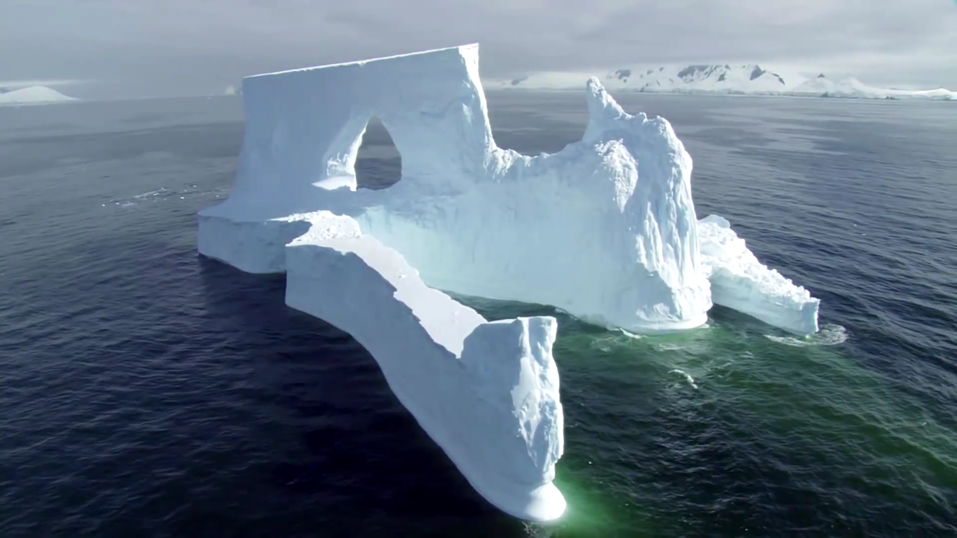 HD PC desktop wallpaper of a majestic iceberg featuring natural arches floating in calm ocean waters under a cloudy sky.