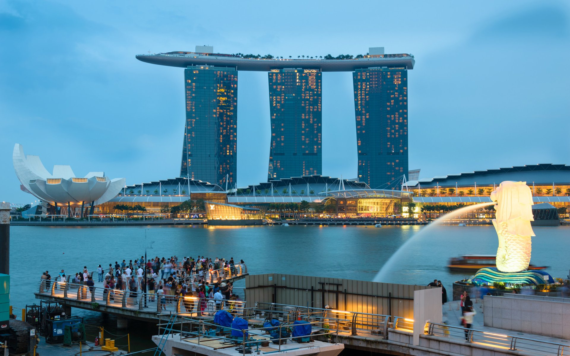 Night view of the Marina Bay Sands building in Singapore with its illuminated structure reflecting on the water, featuring the iconic Merlion statue and nearby crowds.