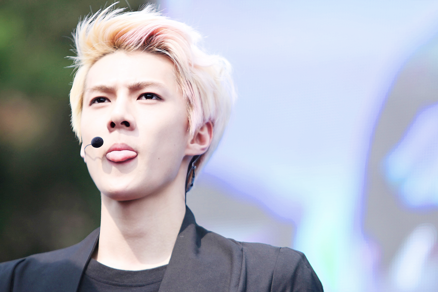 A vibrant HD desktop wallpaper featuring a member of EXO, with a microphone headset, sticking out their tongue against a blurred background. This image captures the energy and style of the music group.