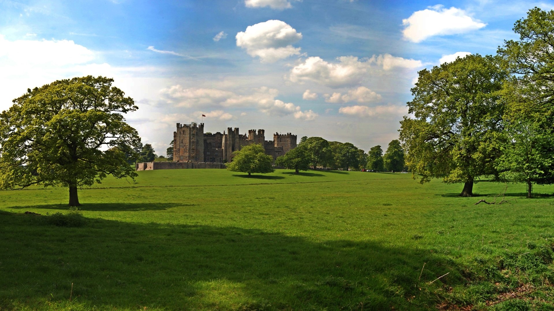 HD PC desktop wallpaper of Raby Castle: man-made medieval castle on expansive green lawns flanked by trees beneath a blue sky with scattered clouds.