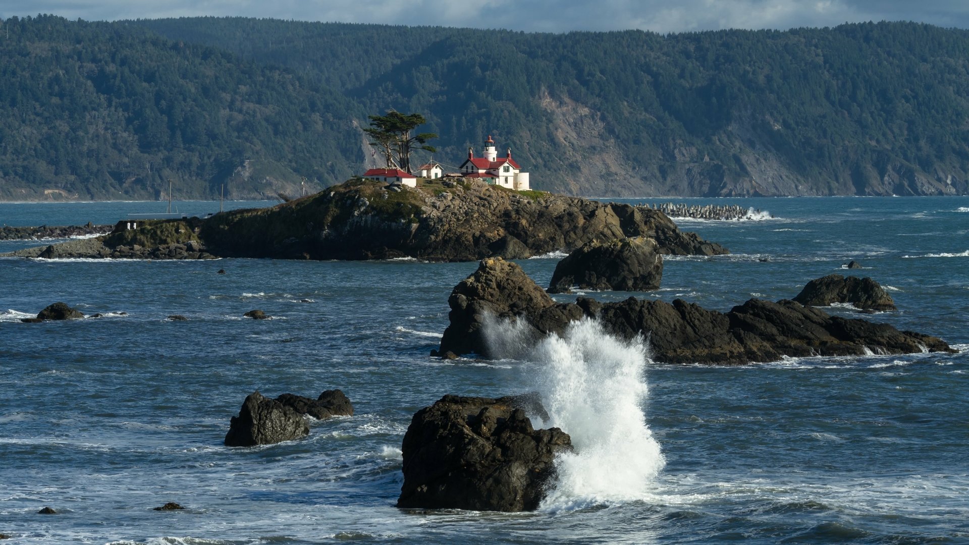 4K Ultra HD wallpaper of a man-made lighthouse on a rocky island with waves crashing in the foreground and forested cliffs in the background.