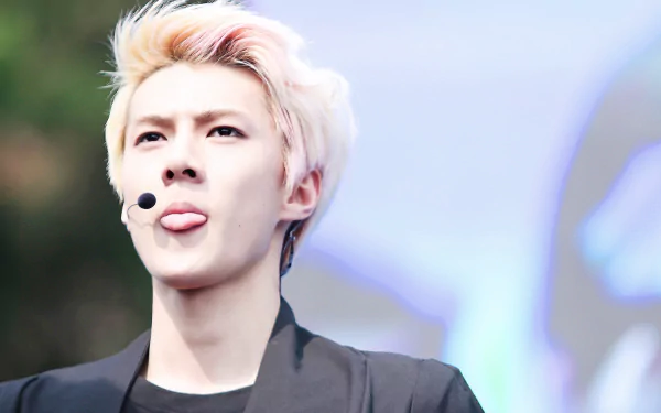 A vibrant HD desktop wallpaper featuring a member of EXO, with a microphone headset, sticking out their tongue against a blurred background. This image captures the energy and style of the music group.