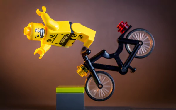 Man-made LEGO figure in bright yellow suit falling off a black bicycle, captured in a dynamic pose with a warm brown background. HD PC desktop wallpaper and background.