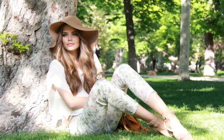HD desktop wallpaper of a woman, Clara Alonso, sitting under a tree in a park, wearing a floppy hat and casual clothing, surrounded by lush greenery.