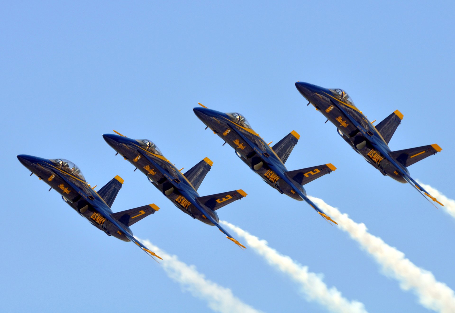 Four McDonnell Douglas F/A-18 Hornet Navy aircraft from the Blue Angels perform a smoke trail formation during a military air show against a clear blue sky.
