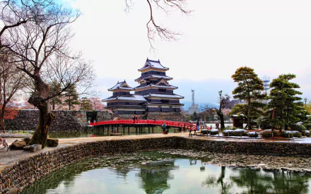 HD PC desktop wallpaper of Matsumoto Castle, a man-made historic Japanese castle with a red arched bridge and moat reflecting the structure, framed by winter trees.