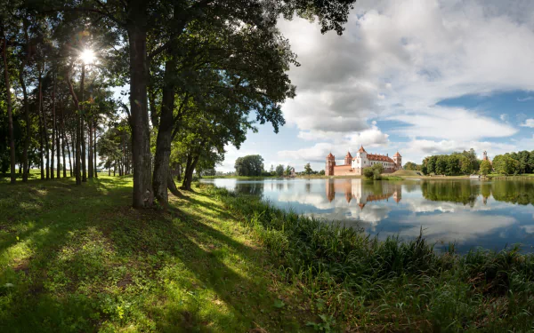 HD desktop wallpaper showcasing Mirsky Castle reflected in calm water, framed by lush trees and a serene sky on a sunny day.