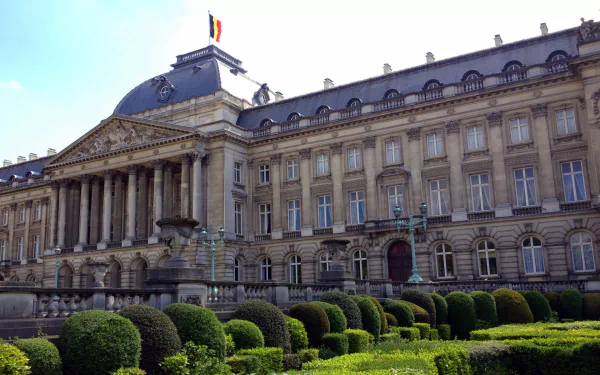 HD desktop wallpaper showing the Royal Palace of Brussels, a grand man-made historic building with manicured gardens and the Belgian flag atop the roof.