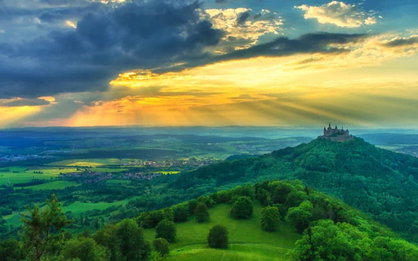 A stunning HD wallpaper showcasing Hohenzollern Castle atop a lush green hill, bathed in golden sunlight with dramatic clouds and a sprawling landscape in the background.