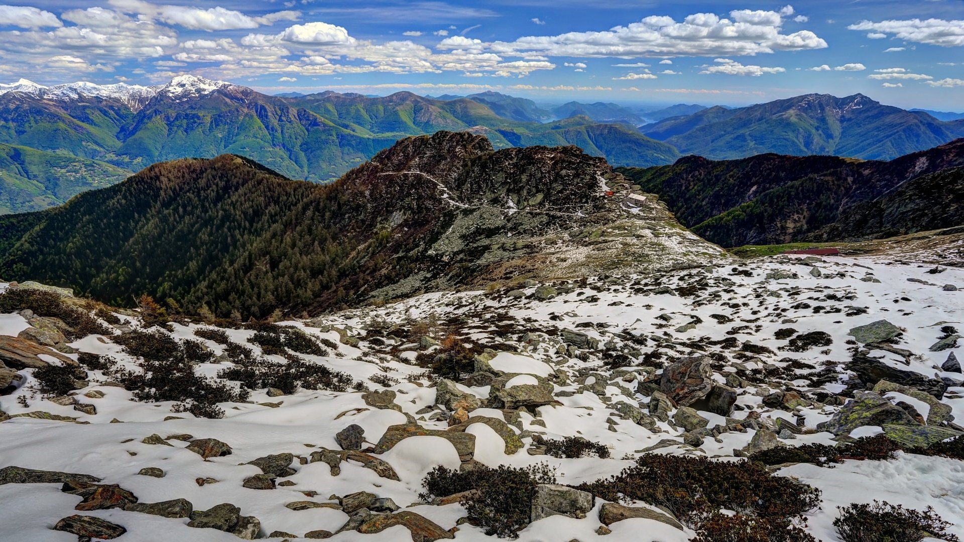 HD PC desktop wallpaper showing a stunning alpine landscape with rocky terrain, patches of snow, and distant mountain peaks under a partly cloudy sky.