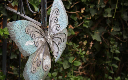 A decorative butterfly sculpture hangs in a lush garden, showcasing intricate designs and a striking blue hue, adding artistic charm to the natural surroundings.