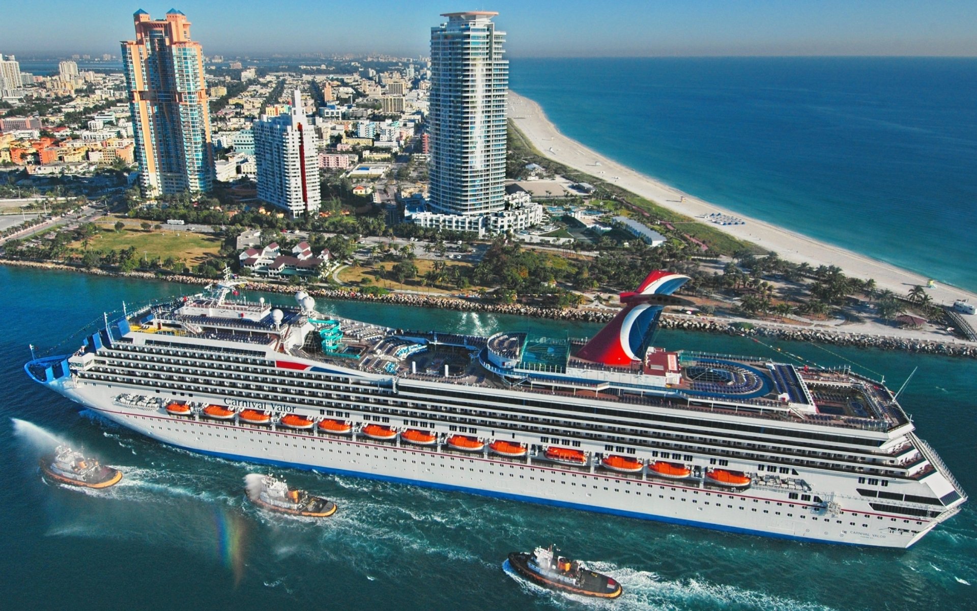 HD desktop wallpaper featuring the Carnival Valor cruise ship sailing near a coastal city with tall buildings and a sandy beach in the background.