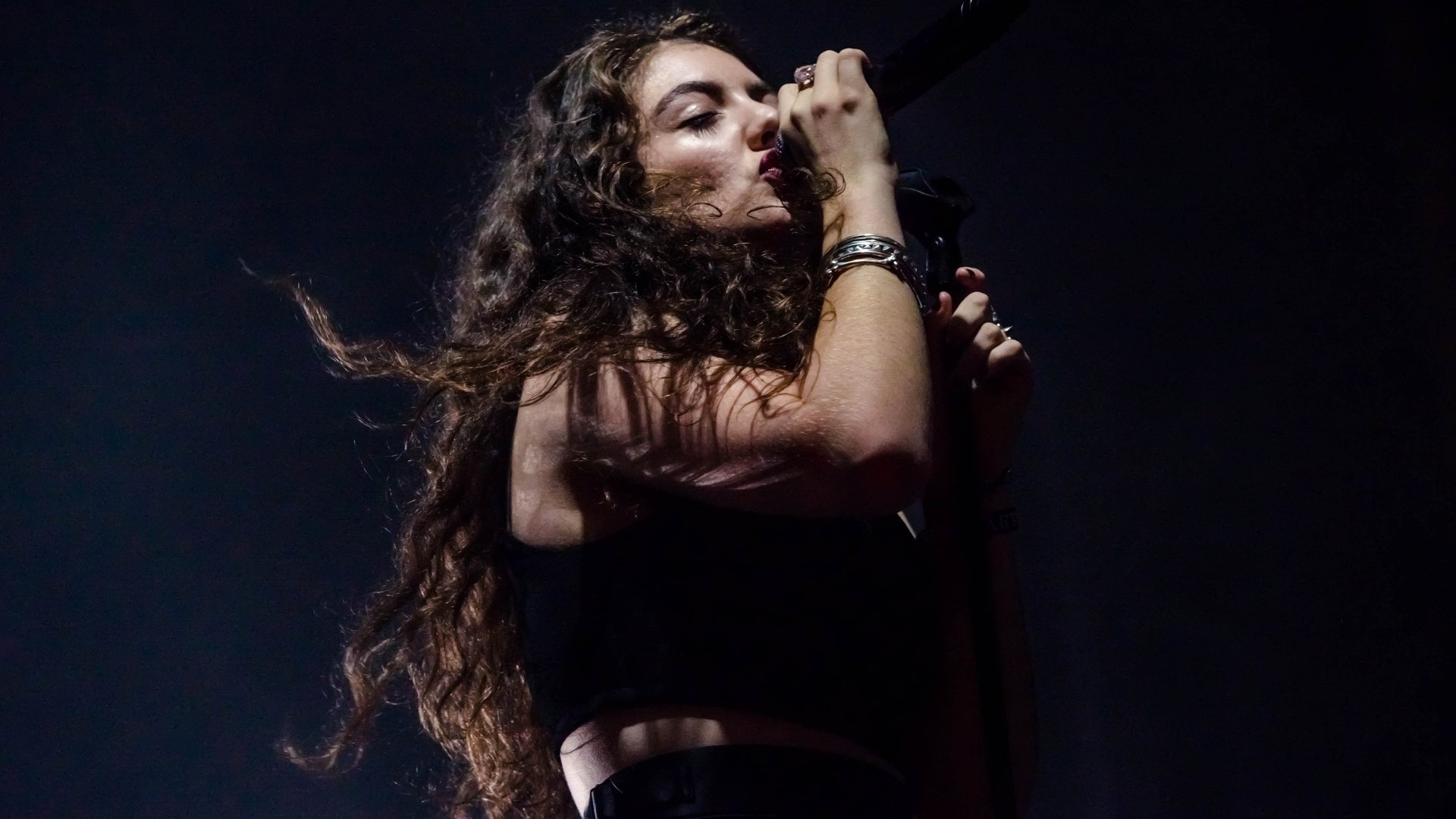 Moody close-up of a female singer mid-performance with long dark hair and mic, deep stage shadows. 5K Ultra HD PC desktop wallpaper/background. Tags: music, Lorde.