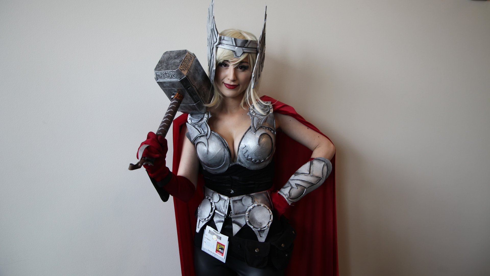A woman in striking cosplay, portraying a heroic character with a hammer, wearing intricate armor and a flowing cape, set against a simple background. High-definition desktop wallpaper.