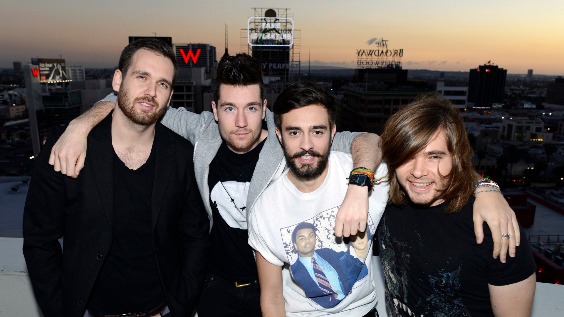 HD desktop wallpaper featuring the English rock band Bastille posing together against a city skyline at sunset.