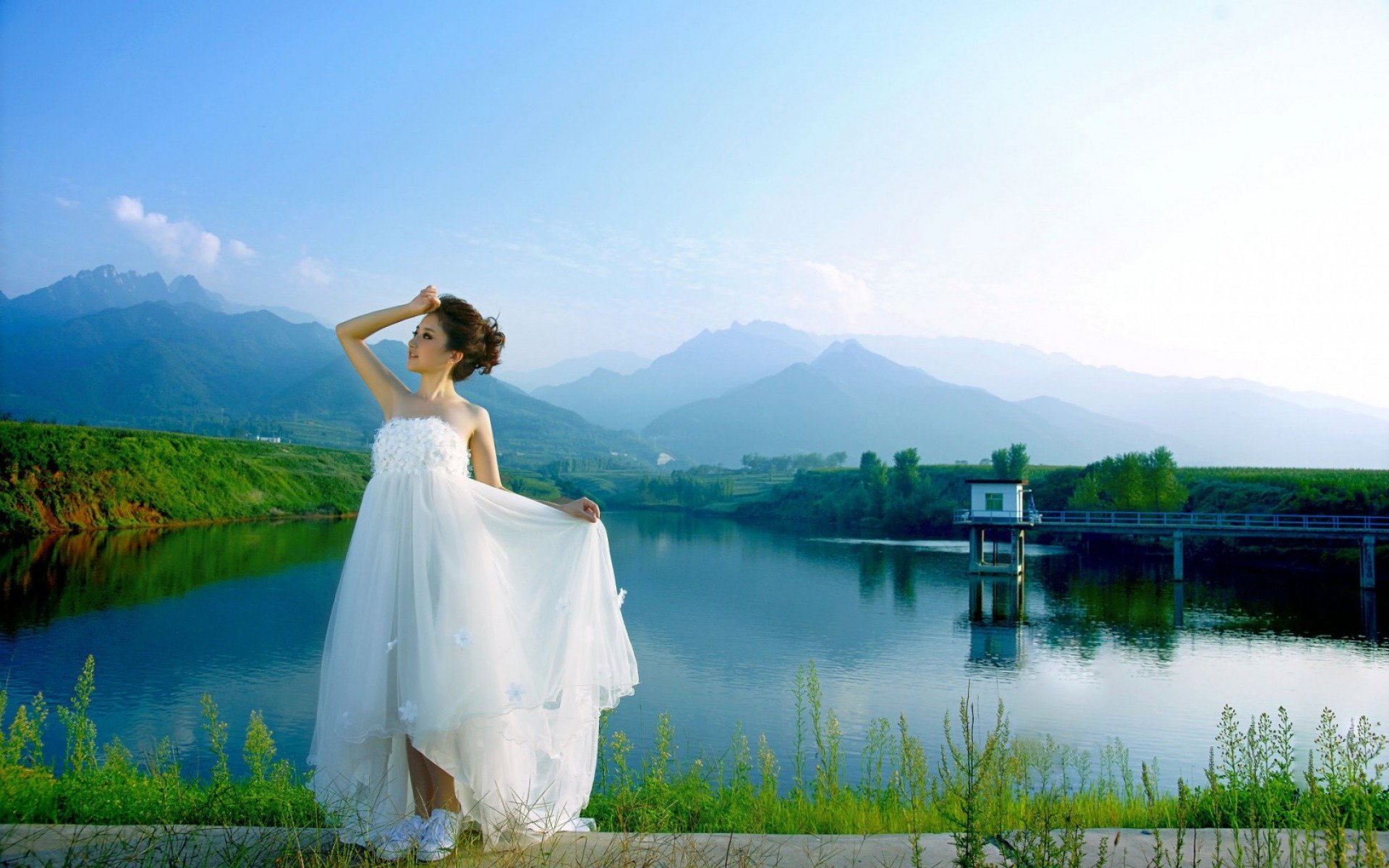 HD desktop wallpaper featuring an Asian woman in a flowing white dress standing by a serene lake with mountains in the background.